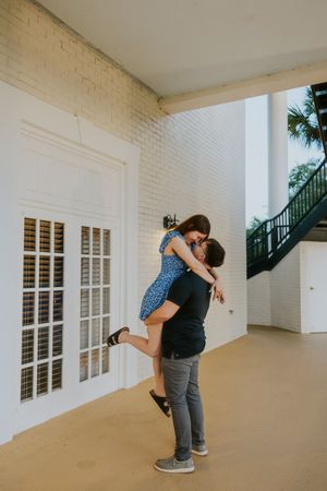 Man lifting woman in blue floral dress in a joyful embrace outside a white brick building.