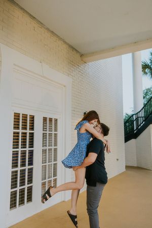 A man lifts a woman in a blue floral dress in a joyful embrace outside a white brick building.