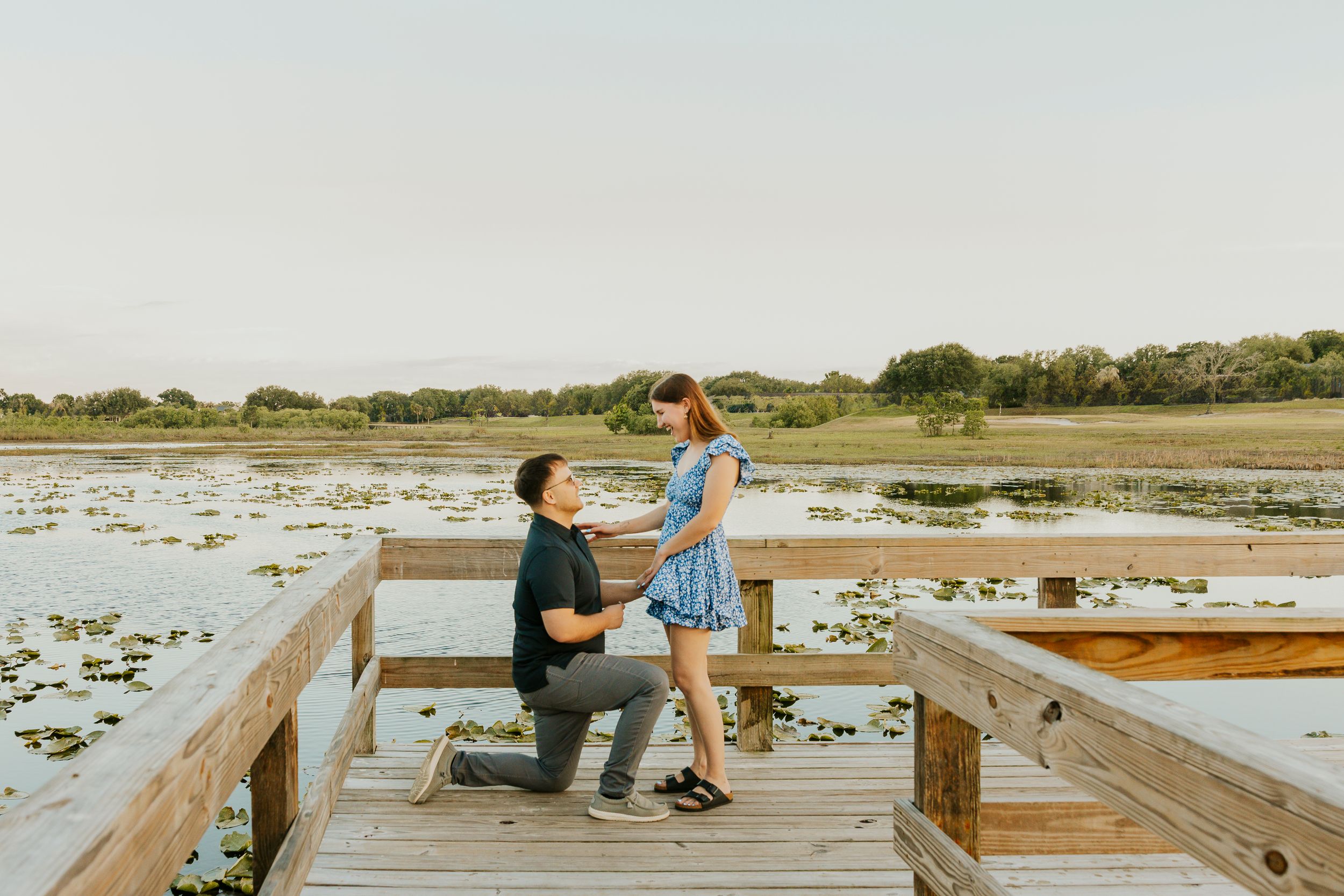 Man proposing on one knee to woman on wooden dock overlooking serene lily pad lake at sunset.
