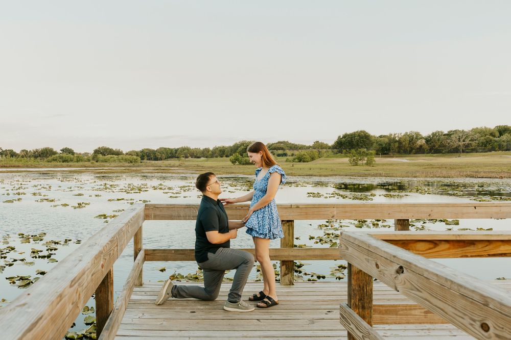 Man proposing on one knee to woman on wooden dock overlooking serene lily pad lake at sunset.