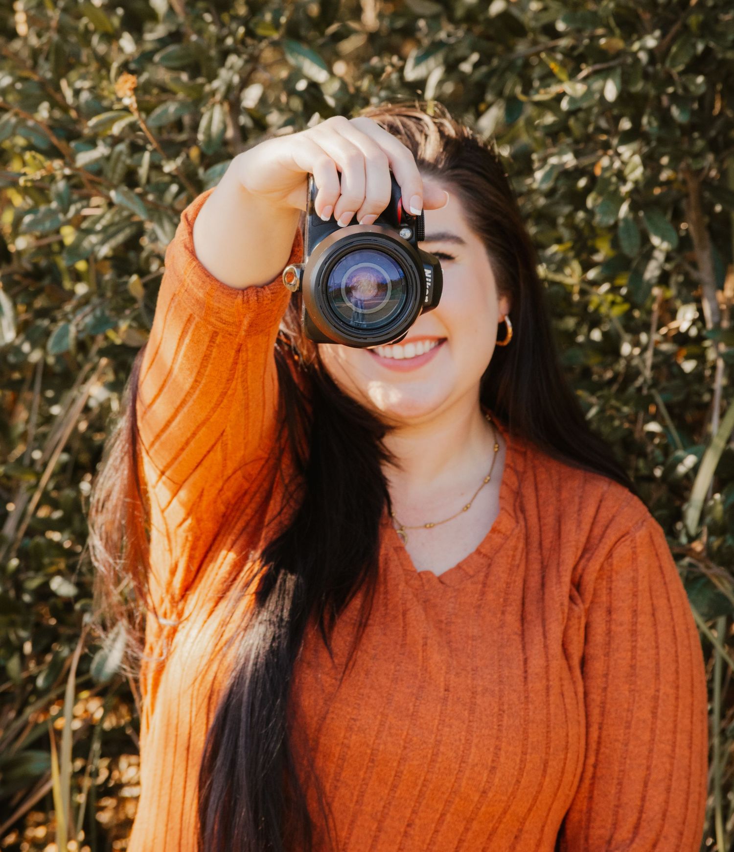Smiling young woman in orange knit sweater holds a Nikon camera up to her face outdoors with green foliage behind her.