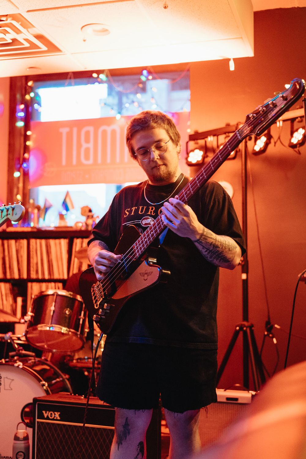 Bassist playing a dark electric bass guitar on stage under warm red lighting at an indoor music venue.