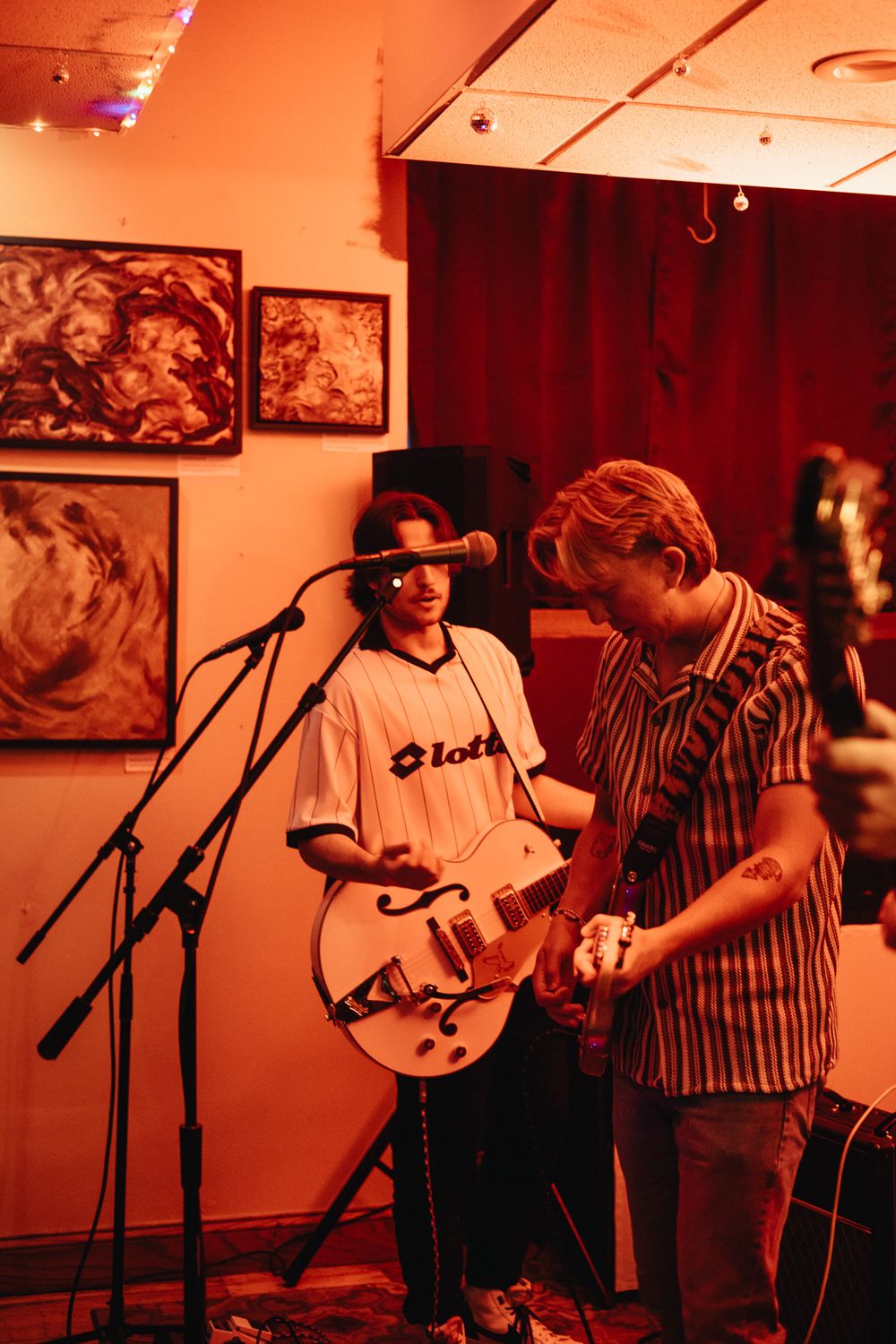 Two musicians playing white guitar on stage under warm red lighting at an indoor music venue.