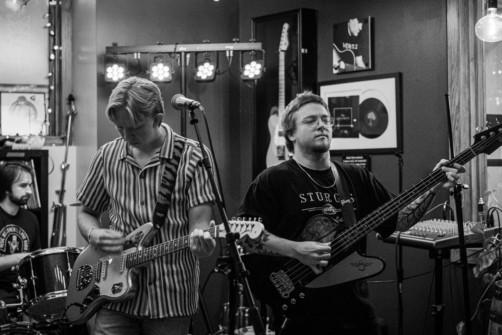 Black and white photo of two guitarists performing live in a music studio with instruments and gold records on the wall.