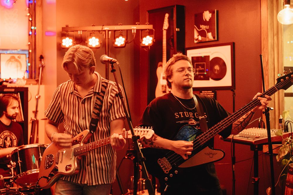 Two musicians playing electric guitars on stage in a warmly lit red room with band memorabilia on the walls.
