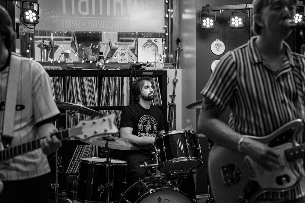 Black and white photo of a band performing in a record store, with a drummer and two guitarists playing live music.