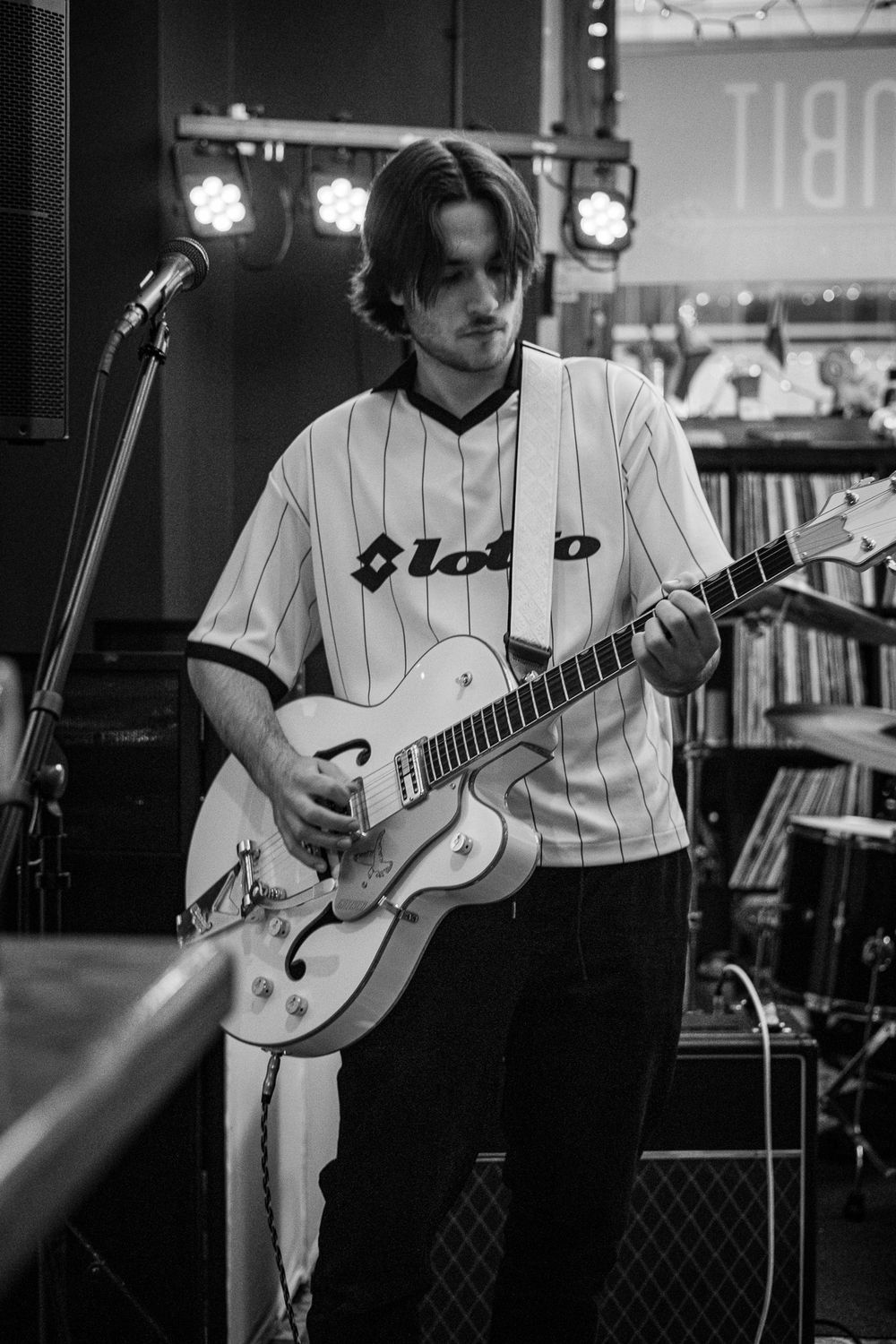 Guitarist in white Lotto baseball jersey plays white hollow-body guitar on stage in black and white photo.