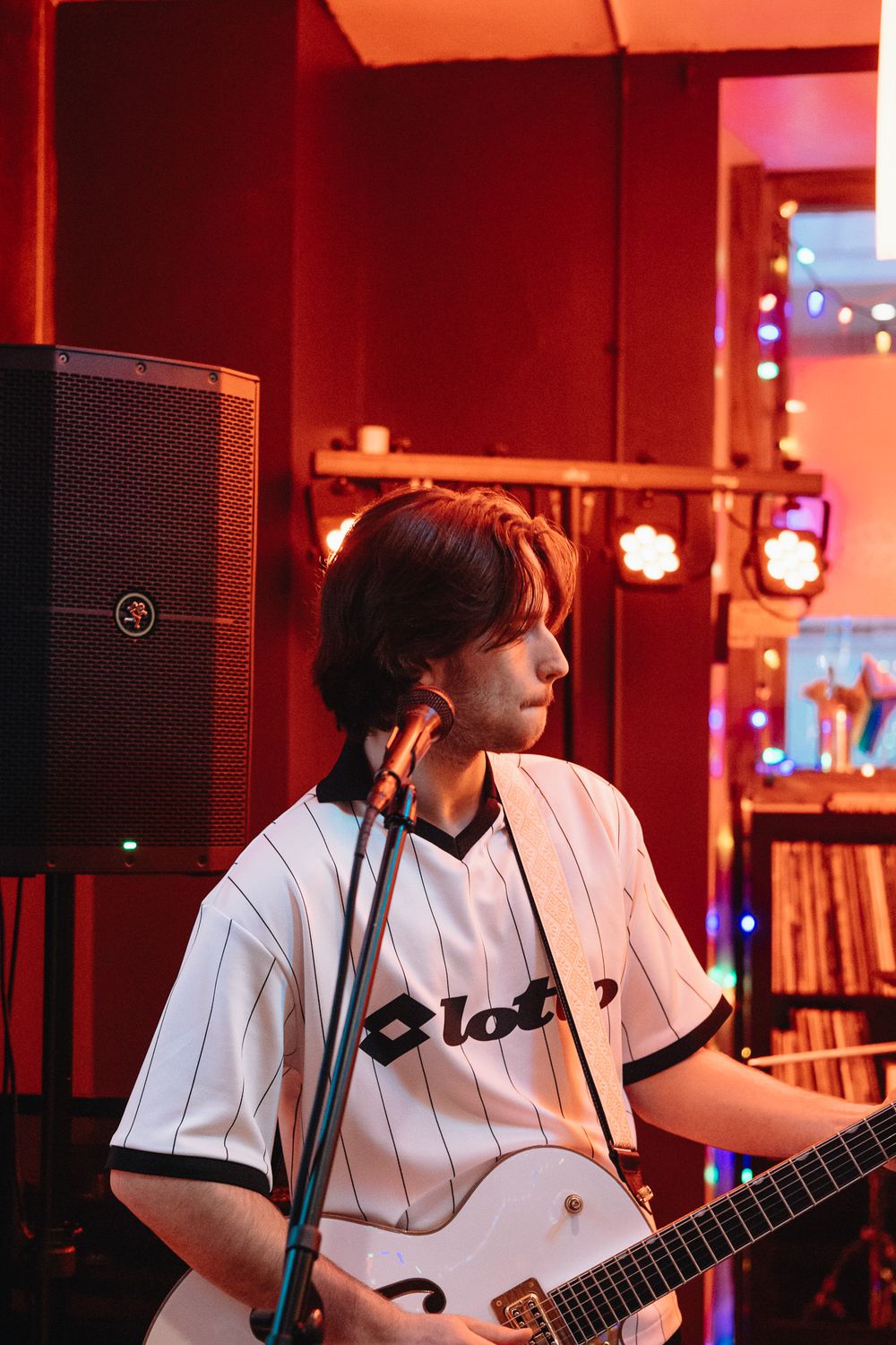 Young guitarist in Lotto jersey performs on stage under red lights at an intimate live music venue.
