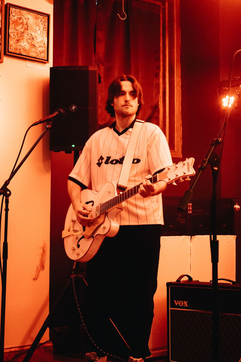 Guitarist in baseball jersey plays white Gretsch guitar on stage under warm red lighting at indoor venue.