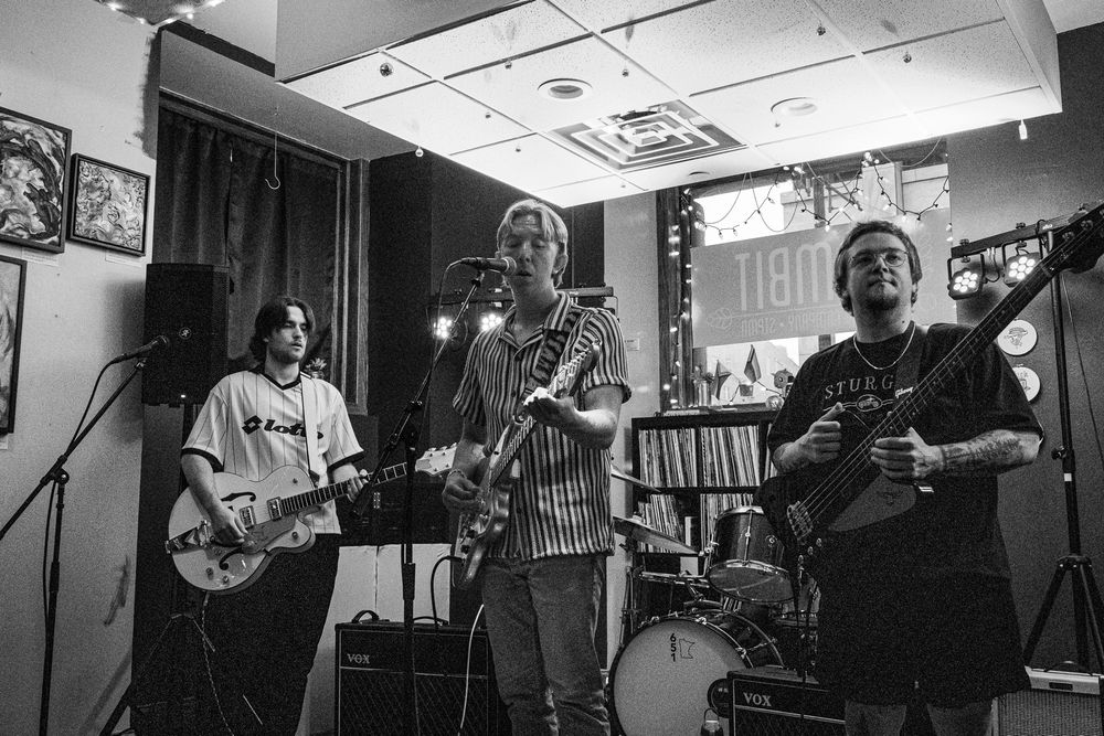 Three-piece rock band performs on small indoor stage in black and white photo at record store.