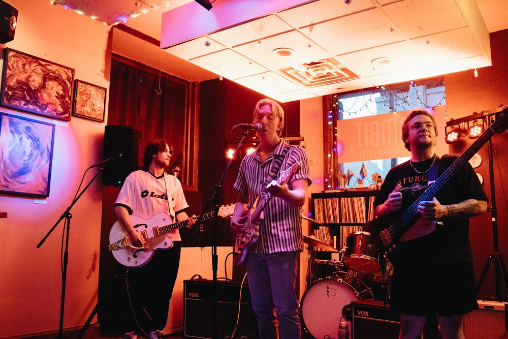 Three musicians perform in a warmly lit venue, two guitarists and a vocalist on stage with drums in the background.