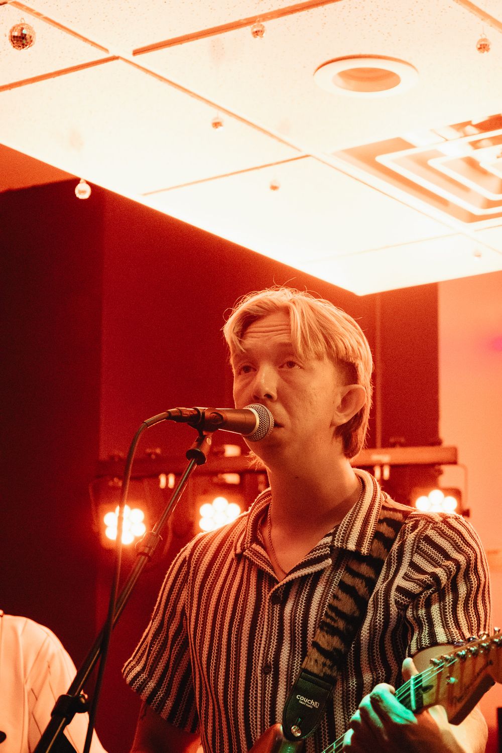 Blonde male vocalist singing into microphone while playing guitar on stage under warm red lighting.