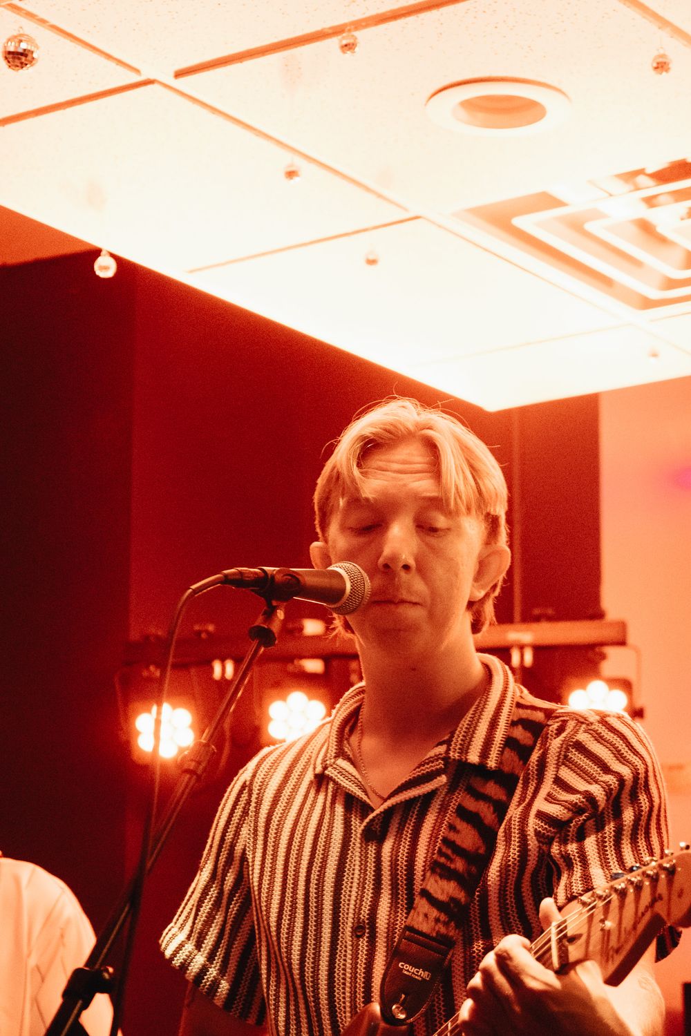 Blonde guitarist sings into microphone wearing striped shirt under warm stage lights at live show.