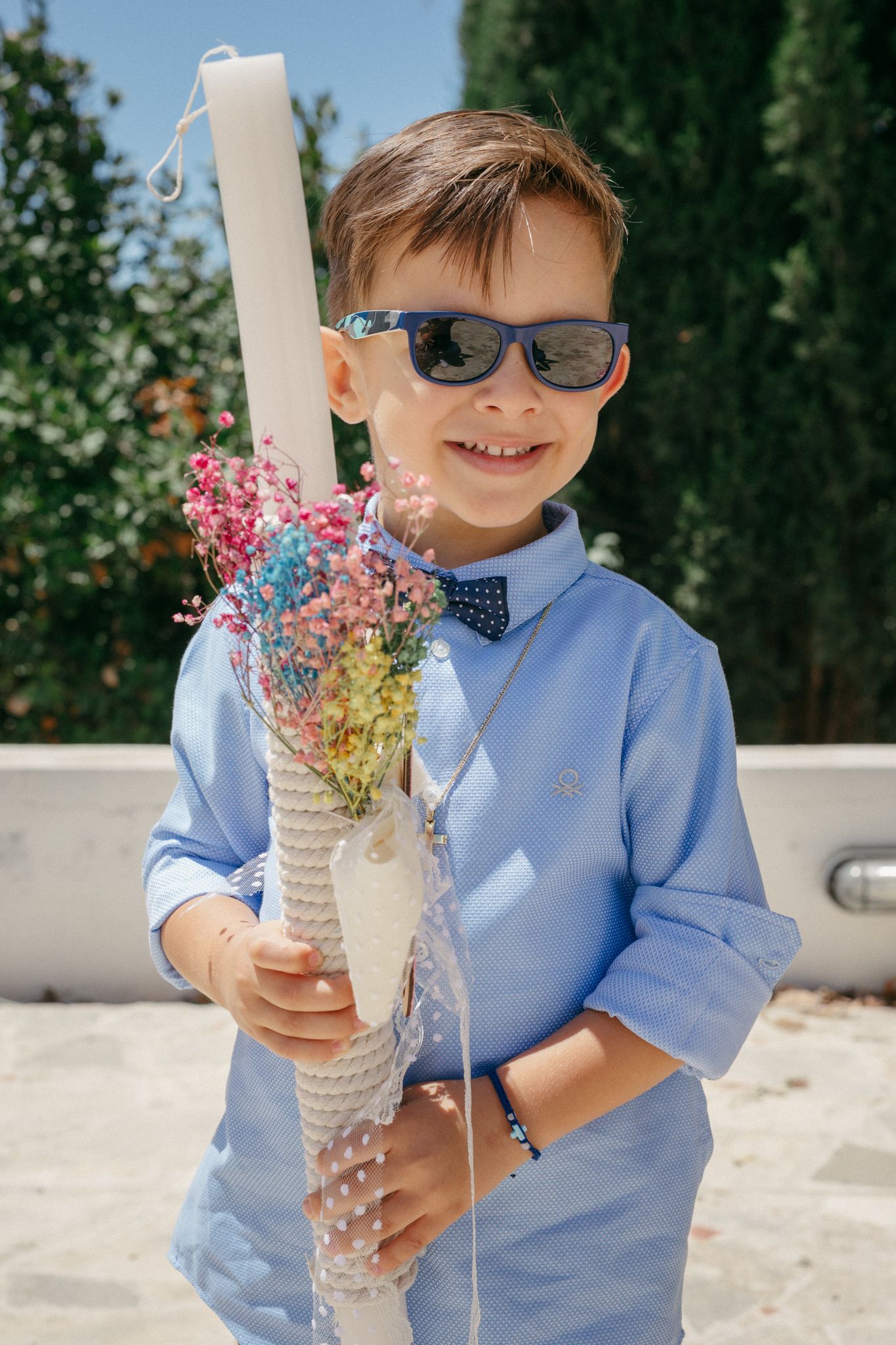 Smiling boy in blue shirt and sunglasses holding a decorated white candle with colorful flowers outdoors.
