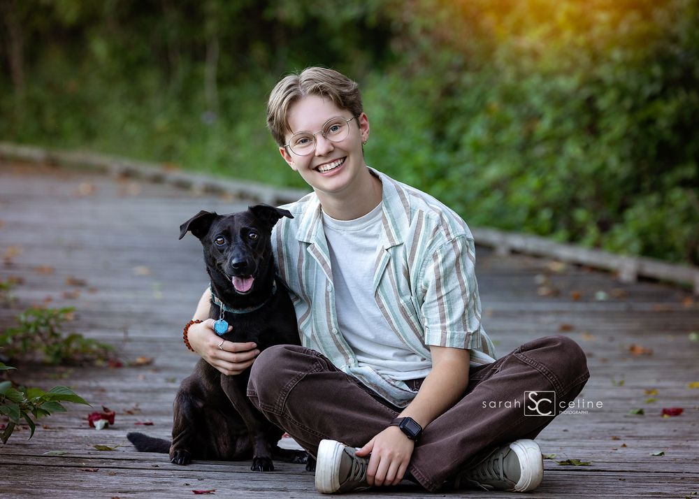 A smiling teen boy sits cross-legged on a wooden deck hugging a black dog outdoors.
