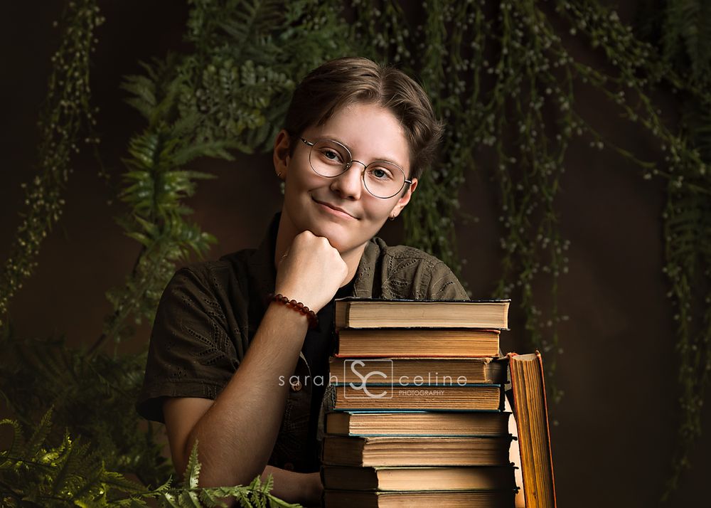 Teen leaning on stack of vintage books with greenery backdrop, wearing glasses and bracelet, smiling warmly at camera.