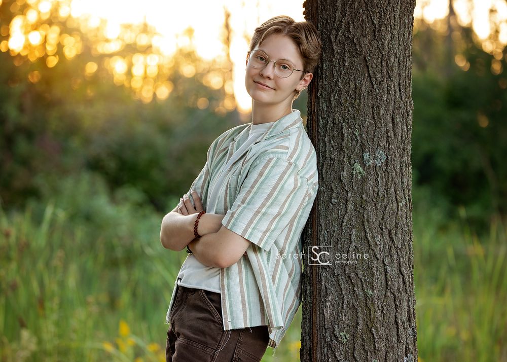 Teen boy with glasses leans against tree in golden hour outdoor portrait session.