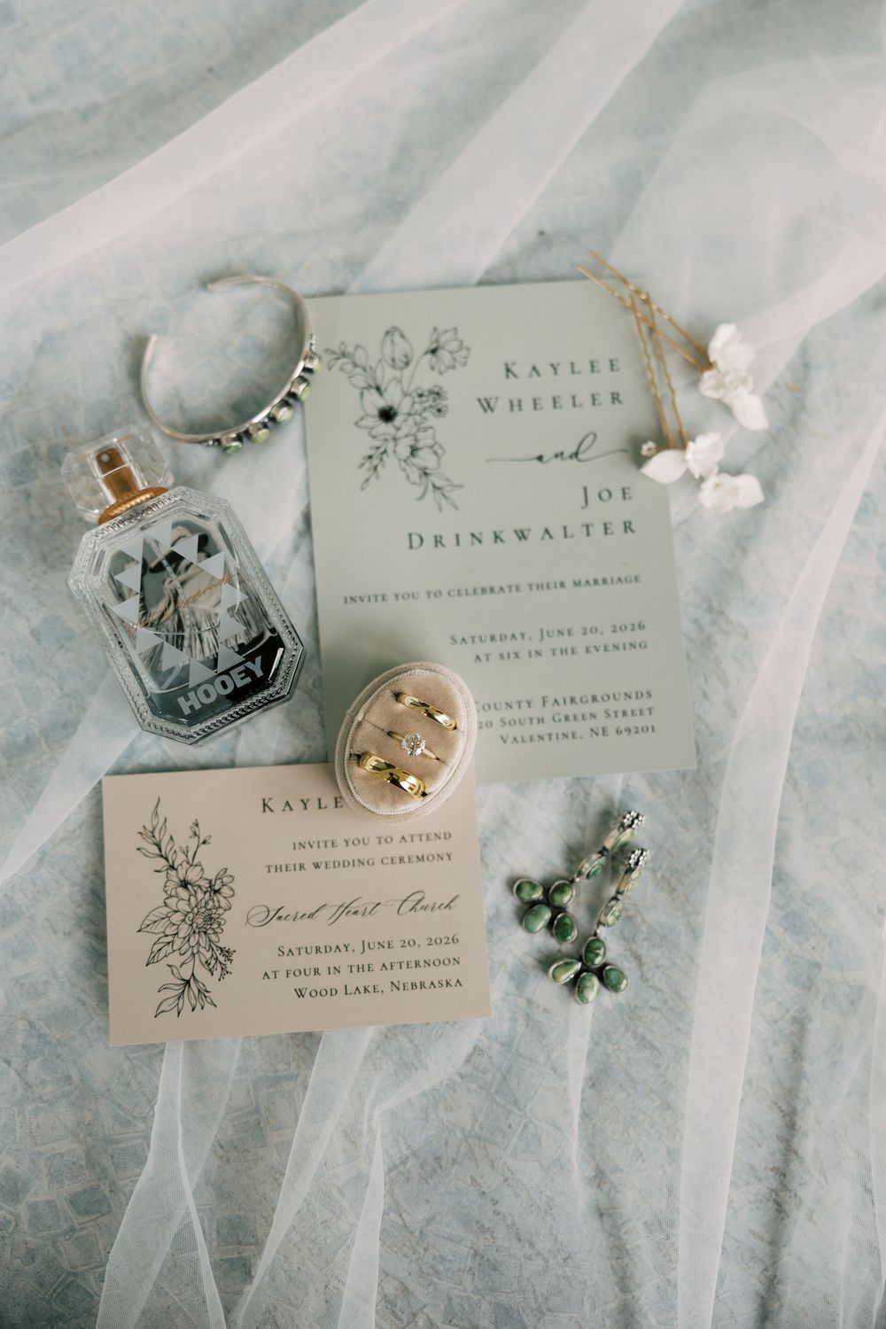 Elegant wedding flat lay featuring invitations for Kaylee Wheeler and Joe Drinkwalter, perfume, jewelry, and accessories on sheer fabric.