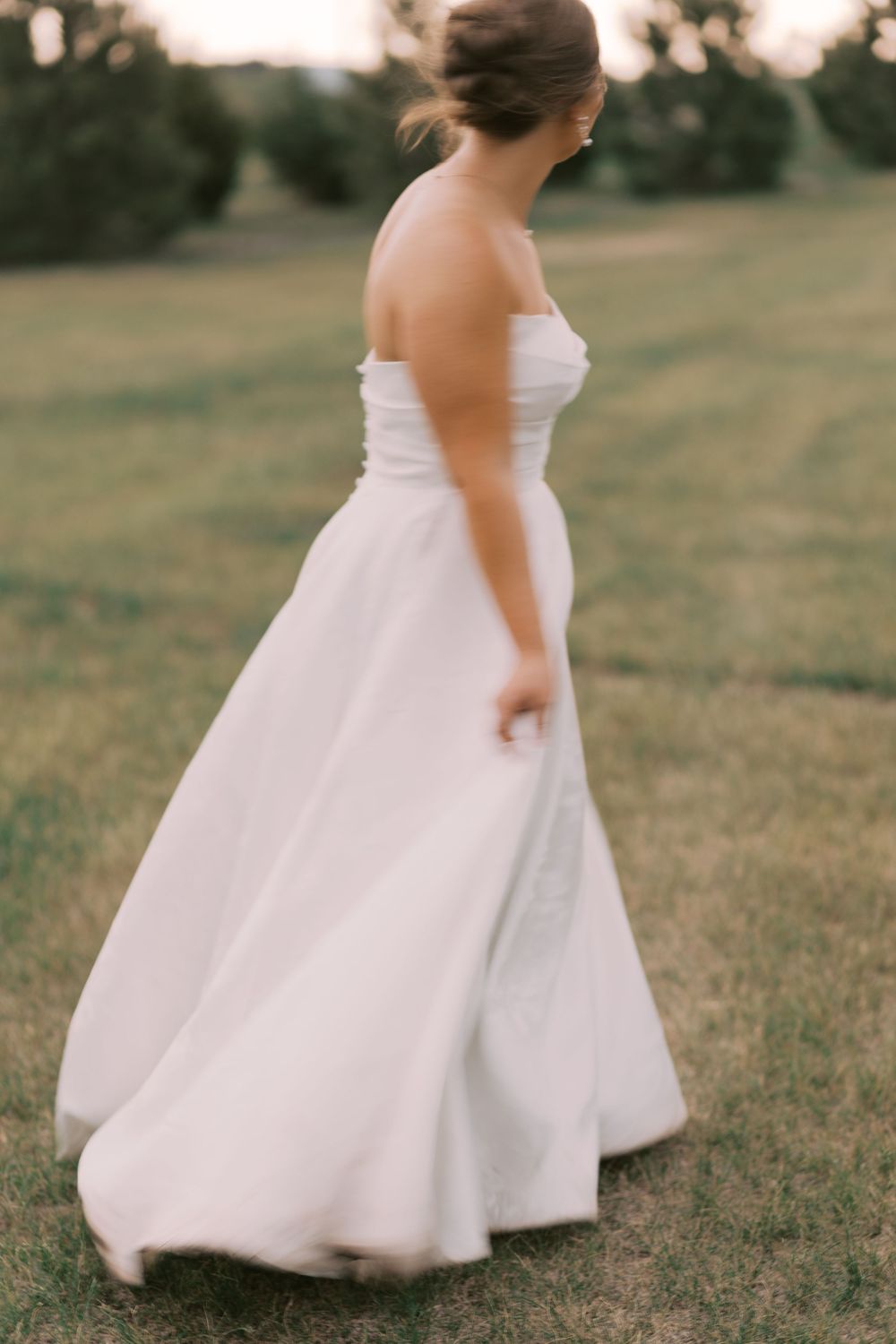 Bride in strapless white ball gown walks gracefully across lush green lawn at golden hour.