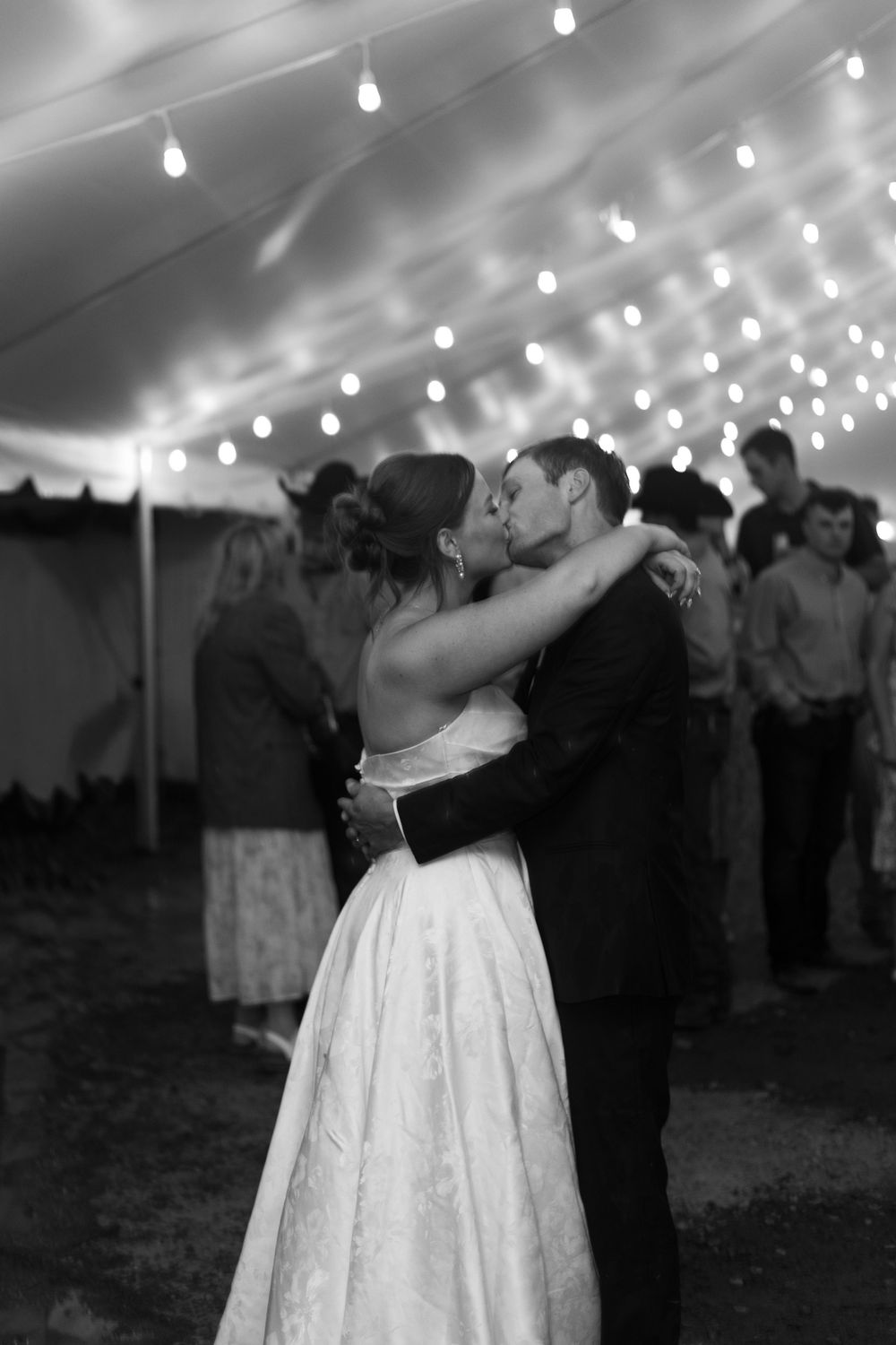 Bride and groom share first dance kiss under twinkling lights in wedding tent, black and white.