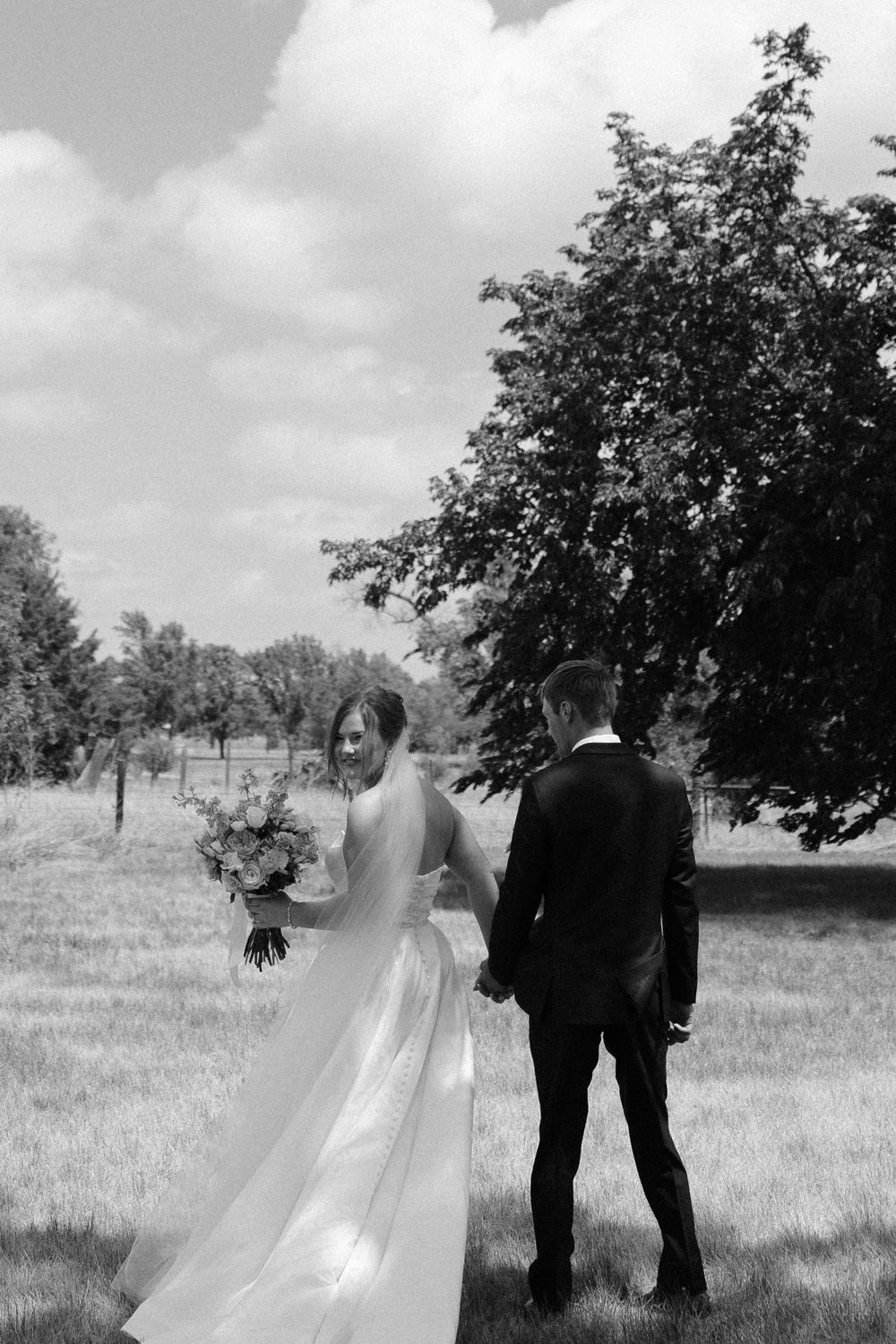 Black and white photo of a bride and groom holding hands walking through a lush green field on their wedding day.