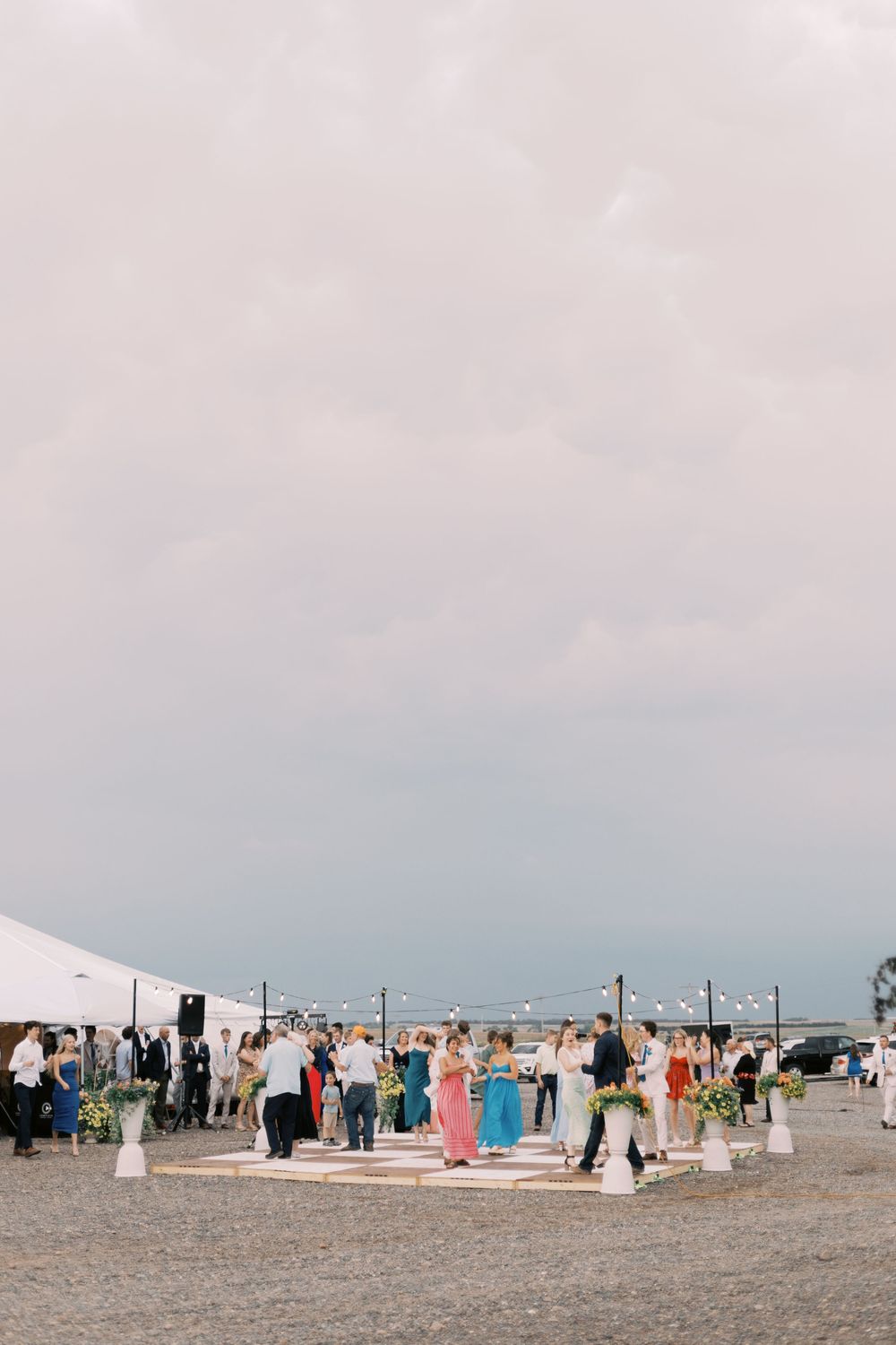 Wedding guests gathered outdoors on cobblestone plaza near white tent under dramatic cloudy sky.