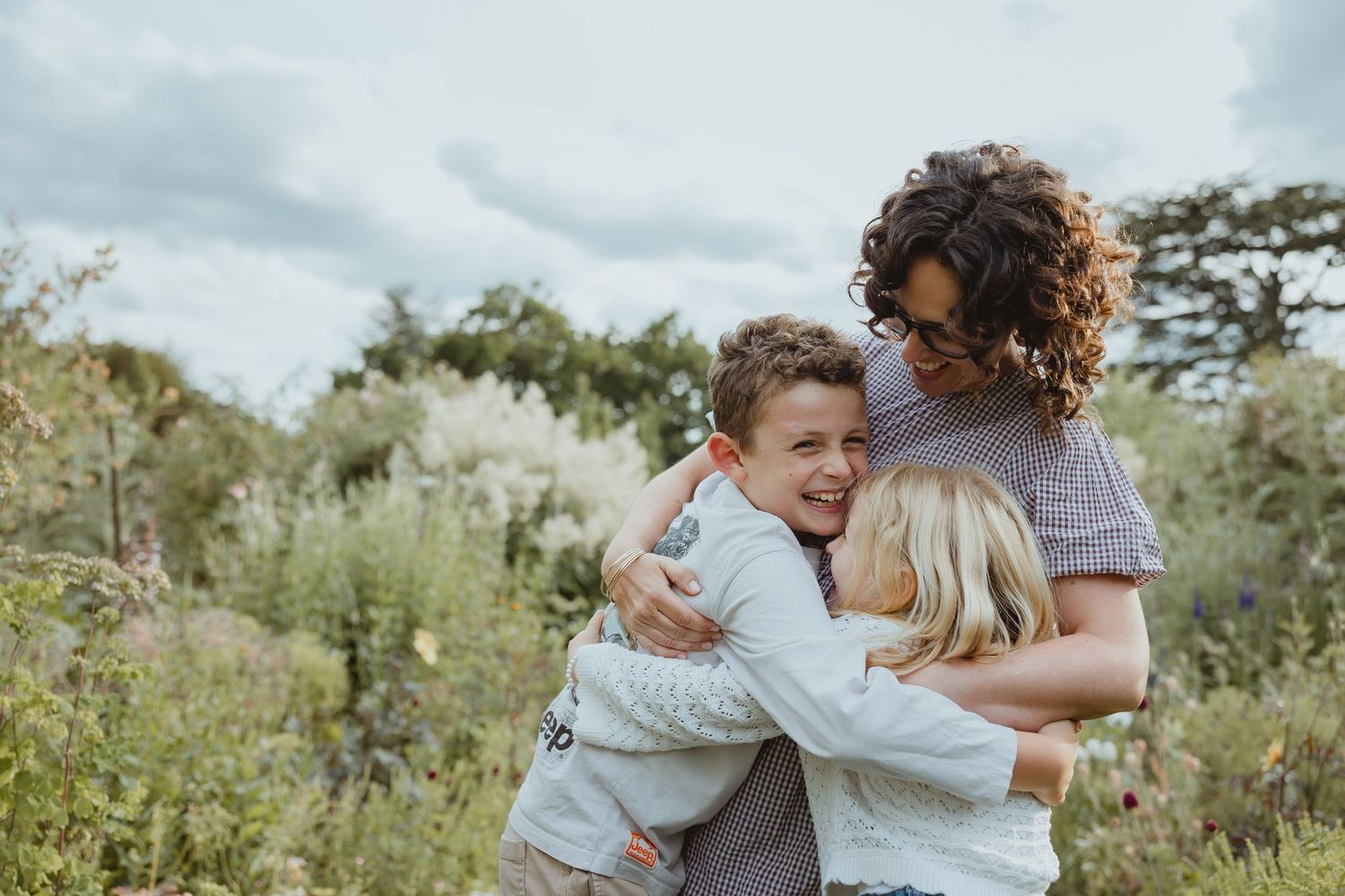 A curly-haired woman hugs two young children in a lush garden setting under a cloudy sky.