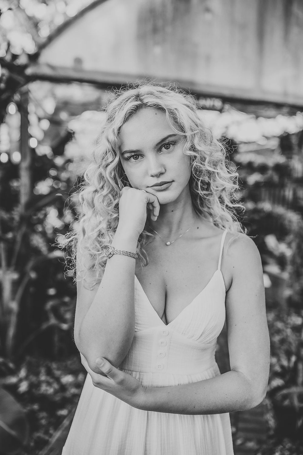 Black and white portrait of a young blonde woman in a white dress posing in a lush garden setting.