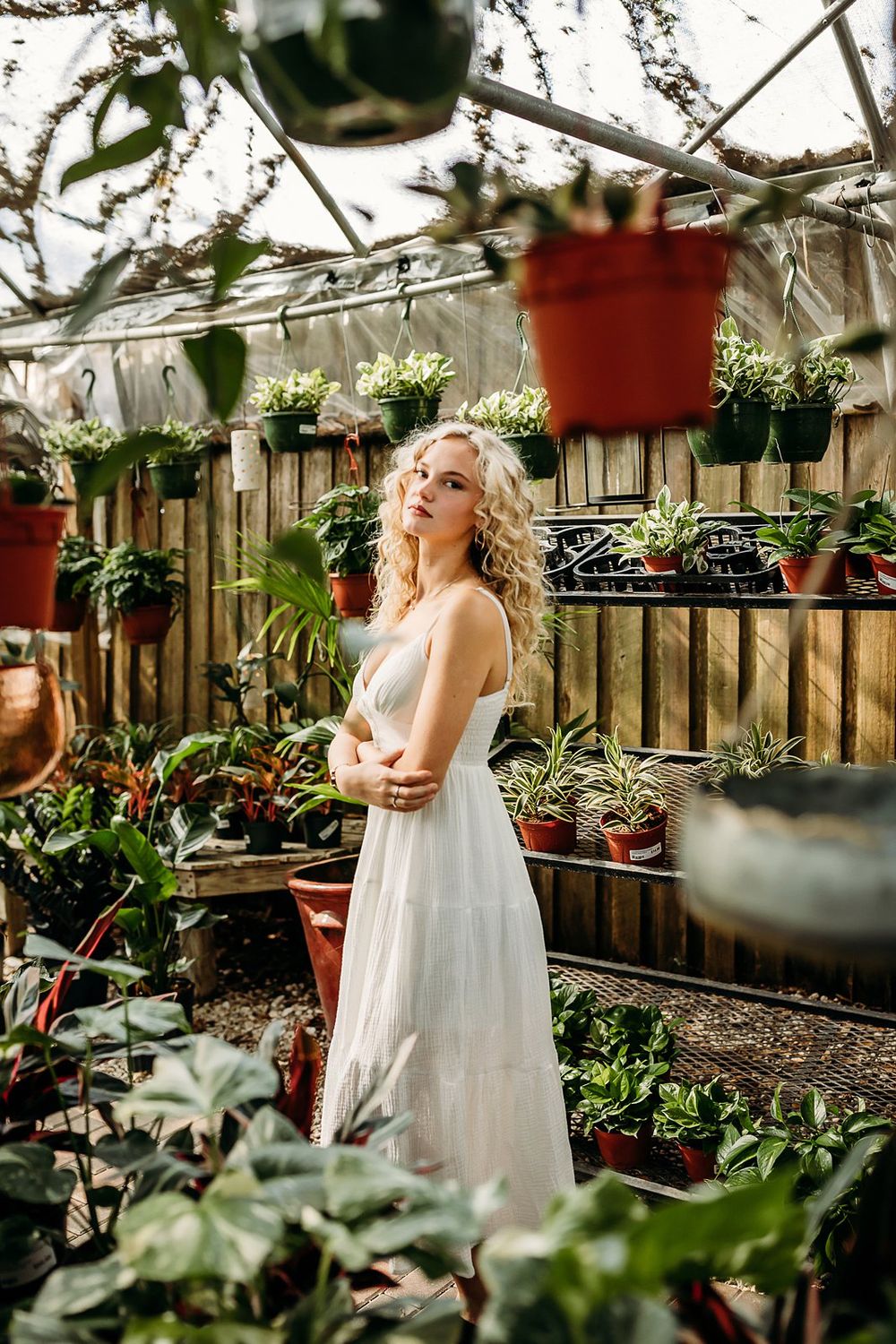 Blonde woman in white dress posing gracefully among lush potted plants in a sunlit greenhouse.