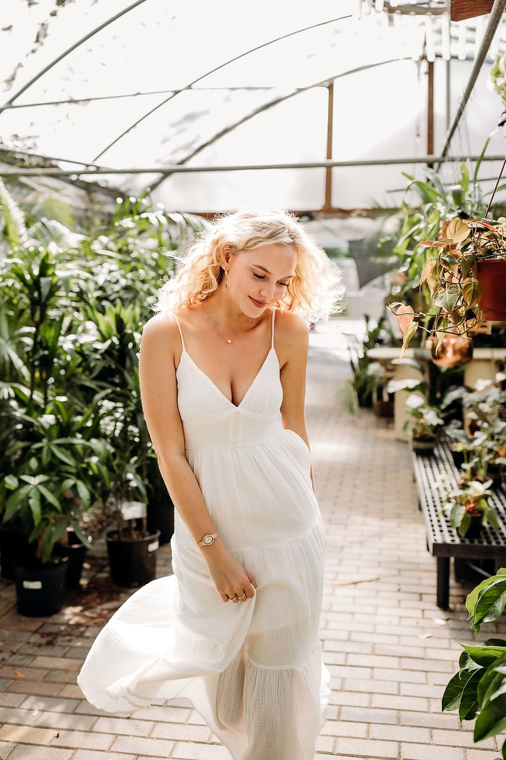 Blonde woman in white spaghetti strap dress posing gracefully in a lush greenhouse surrounded by tropical plants.