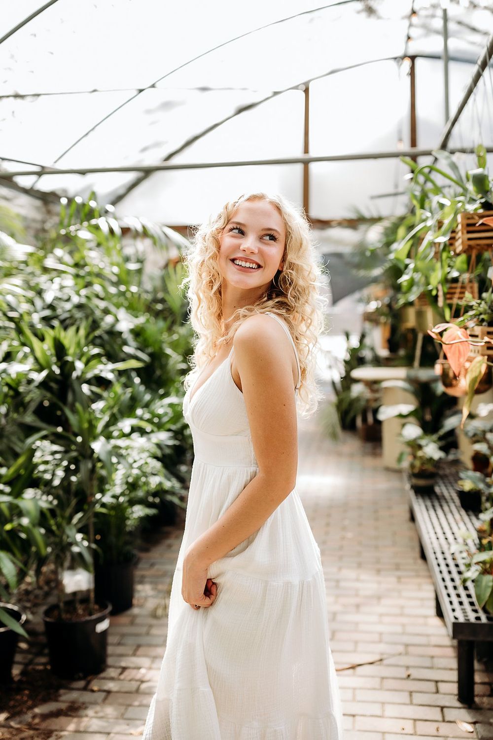 Smiling blonde woman in white dress standing in a lush greenhouse surrounded by tropical plants.