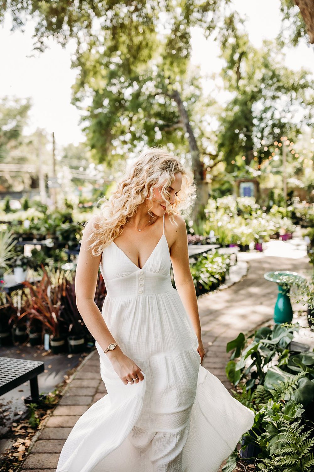 Blonde woman in white tiered spaghetti strap wedding dress walking through lush garden nursery.
