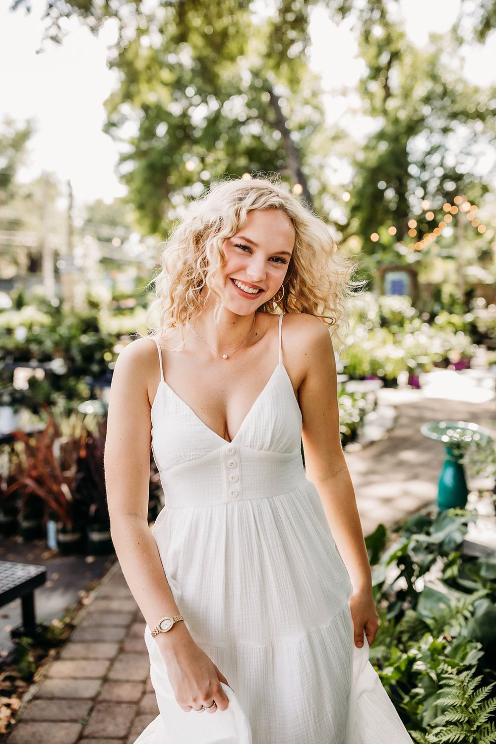Smiling blonde woman in a white sundress standing in a sunny outdoor garden with lush greenery.