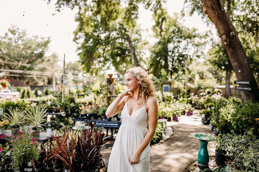Blonde woman in white dress standing among lush garden plants and flowers at an outdoor nursery.