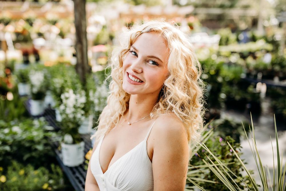 Smiling blonde woman with curly hair in a white dress surrounded by lush green plants in a garden center.