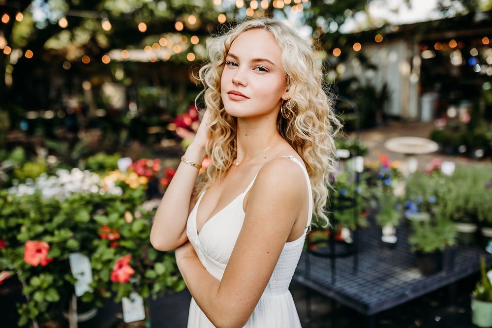 Young blonde woman in white dress poses among colorful flowers and string lights outdoors.