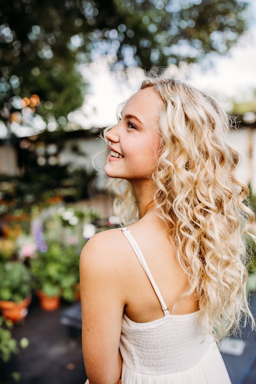 Young blonde woman with curly hair smiling outdoors in a white sundress surrounded by lush green plants.