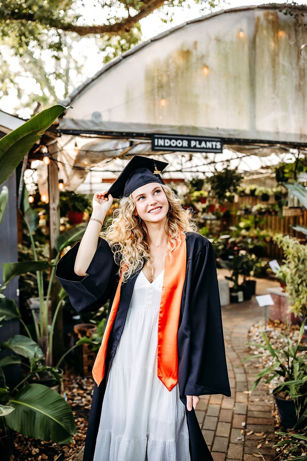 Smiling graduate in black cap and gown with orange sash poses among lush plants at an indoor plant nursery.