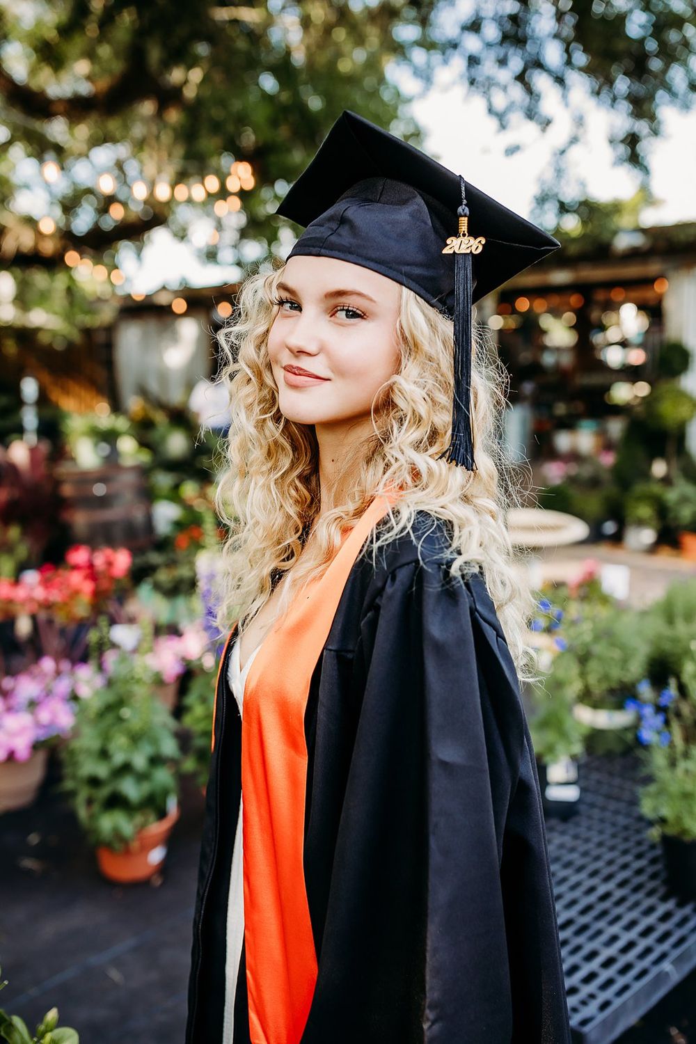 Smiling blonde graduate in black cap and gown with orange sash posing outdoors among colorful flowers in 2020.