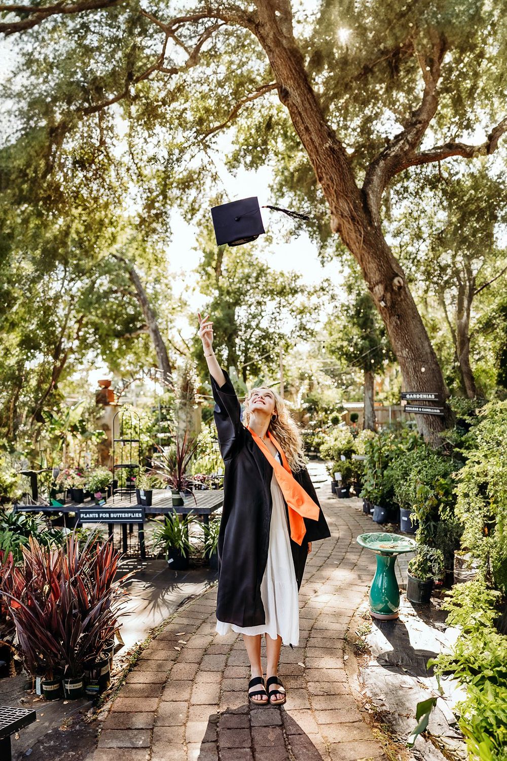 Blonde graduate in black gown tosses cap in lush garden path, celebrating academic achievement outdoors.