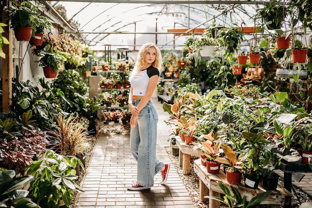 Blonde woman in jeans and white top posing in a lush greenhouse surrounded by tropical plants and flowers.