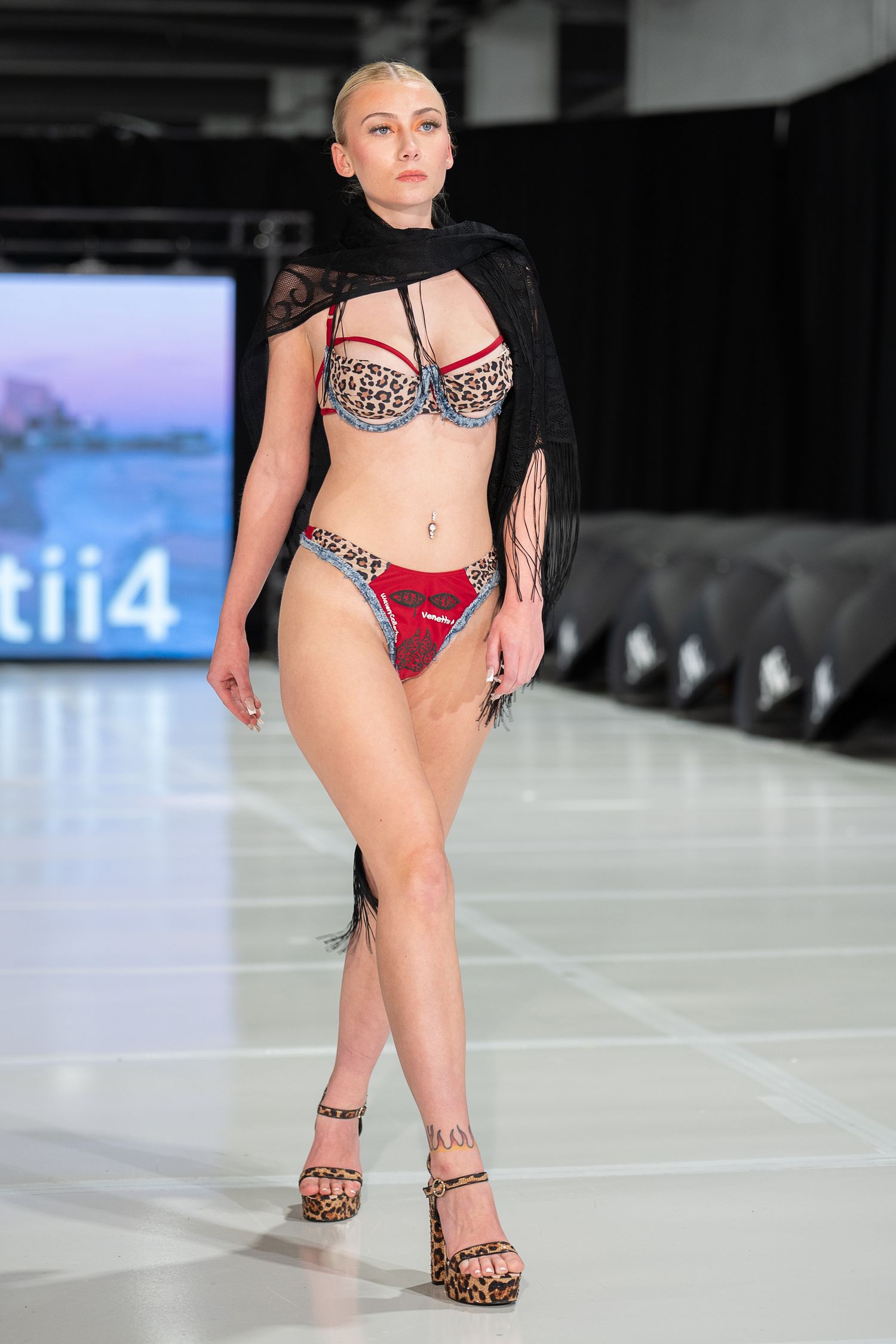 Model walks runway in leopard print bikini with red accents and black fringe wrap at a fashion show.