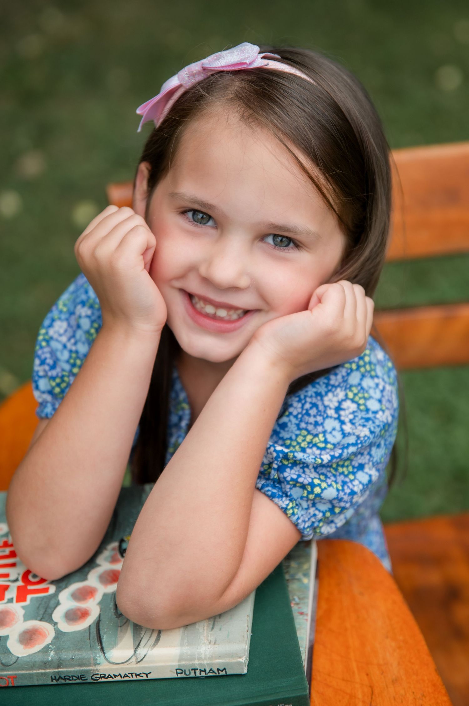 Smiling young girl with pink bow resting chin on hands over books outdoors in blue floral dress.