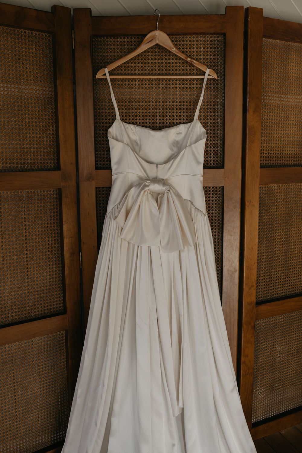 Elegant ivory satin wedding gown with bow detail hanging on wooden hanger against rattan screen.