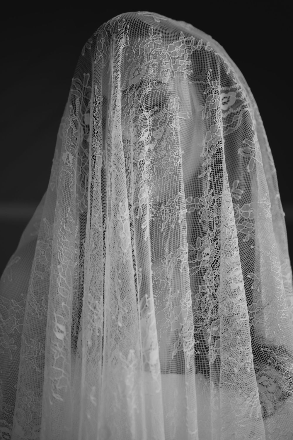 Black and white photo of a person draped in an intricate white lace bridal veil covering their face.