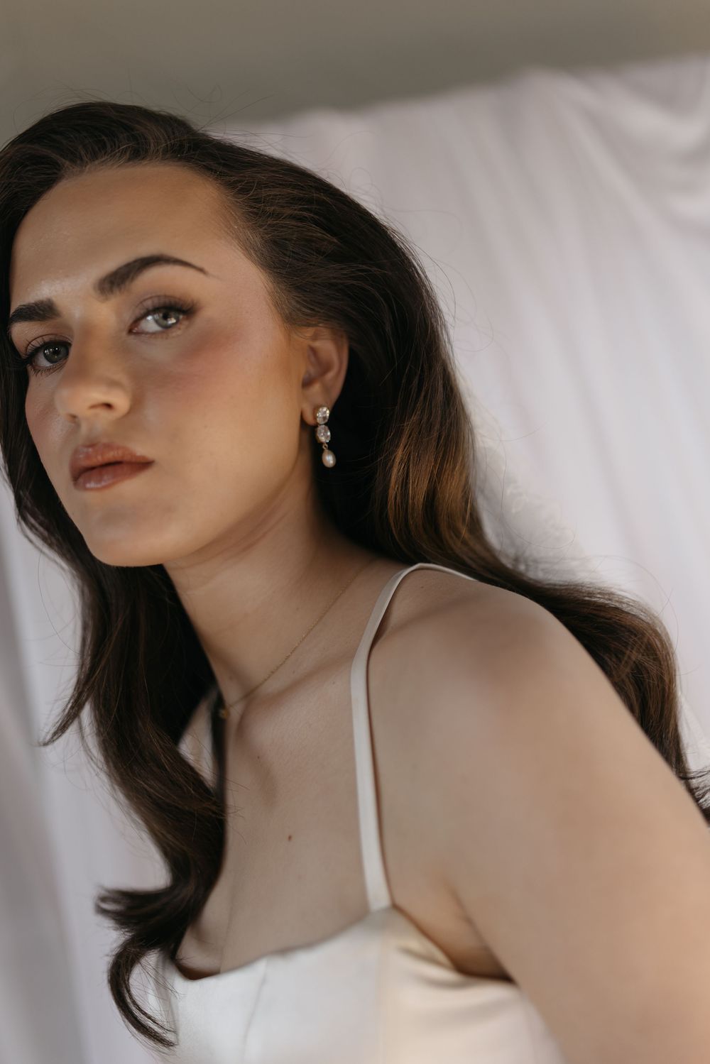Brunette bride wearing delicate drop earrings and gold necklace in white satin wedding dress.