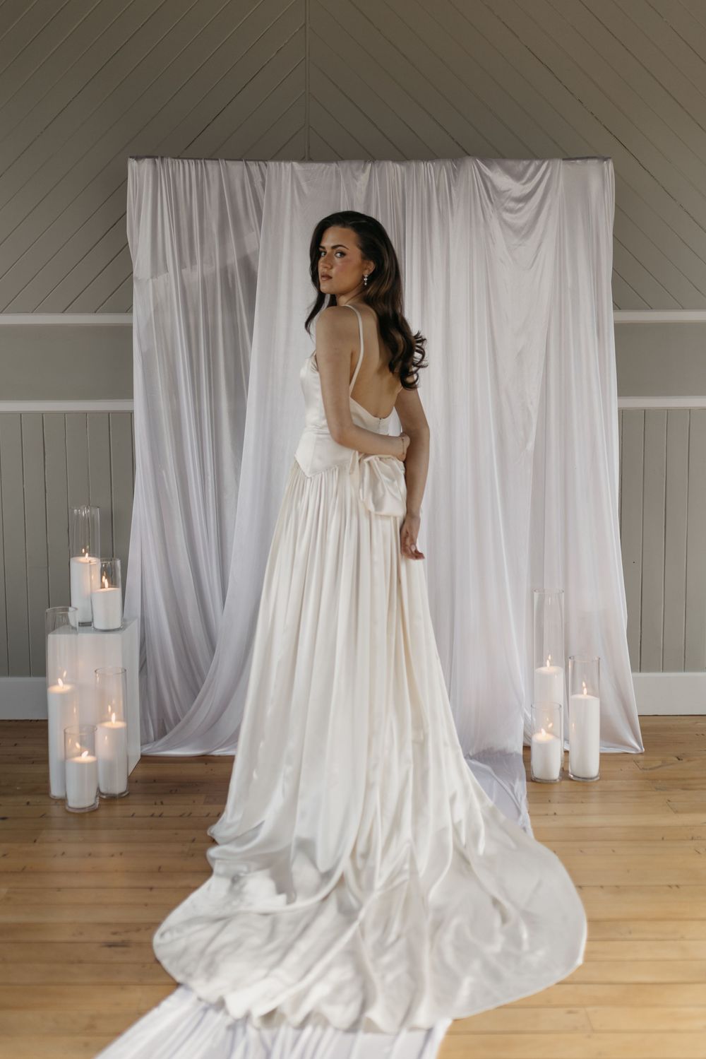 Bride in elegant white satin backless wedding gown with train, posing before draped fabric backdrop with candles.