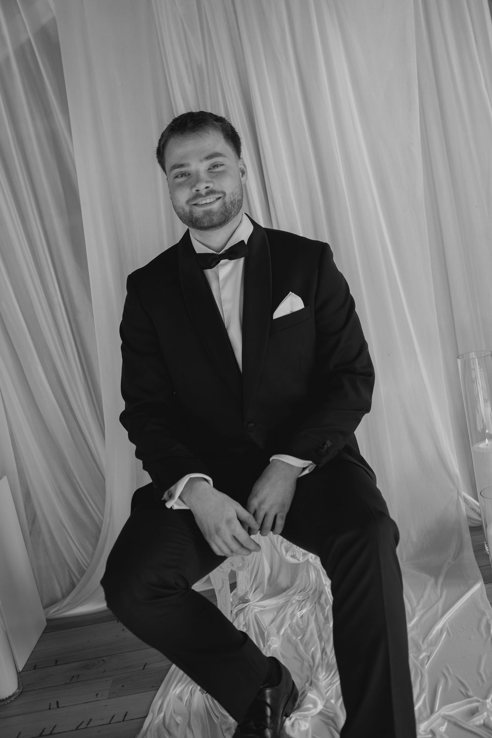Handsome groom in classic black tuxedo with bow tie and pocket square, seated and smiling in black and white photo.