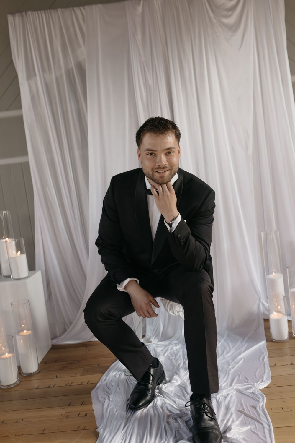 A well-dressed man in a black tuxedo sits confidently before a white draped backdrop surrounded by pillar candles.