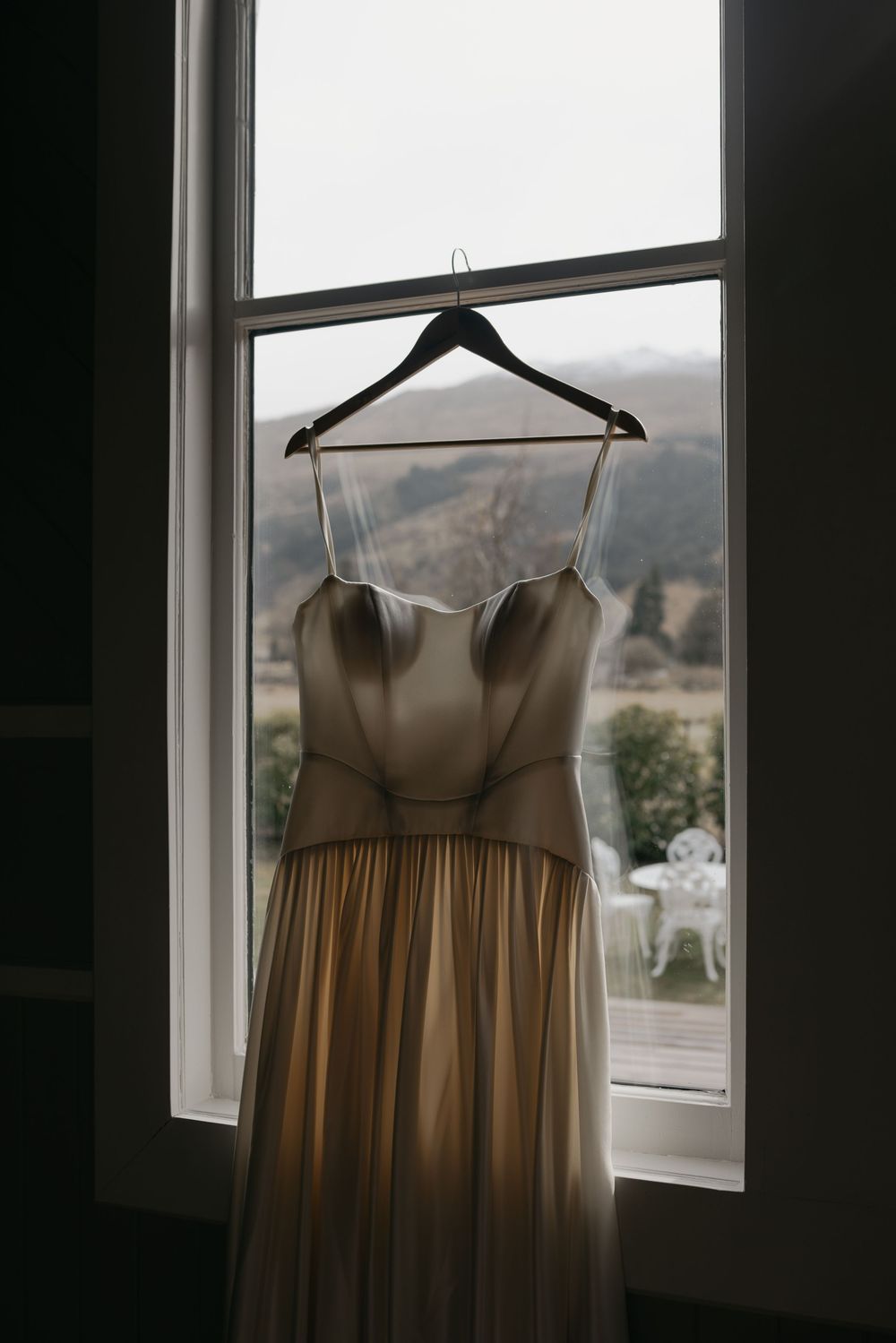 Elegant gold satin wedding dress hanging on a hanger in a window with a scenic countryside view.